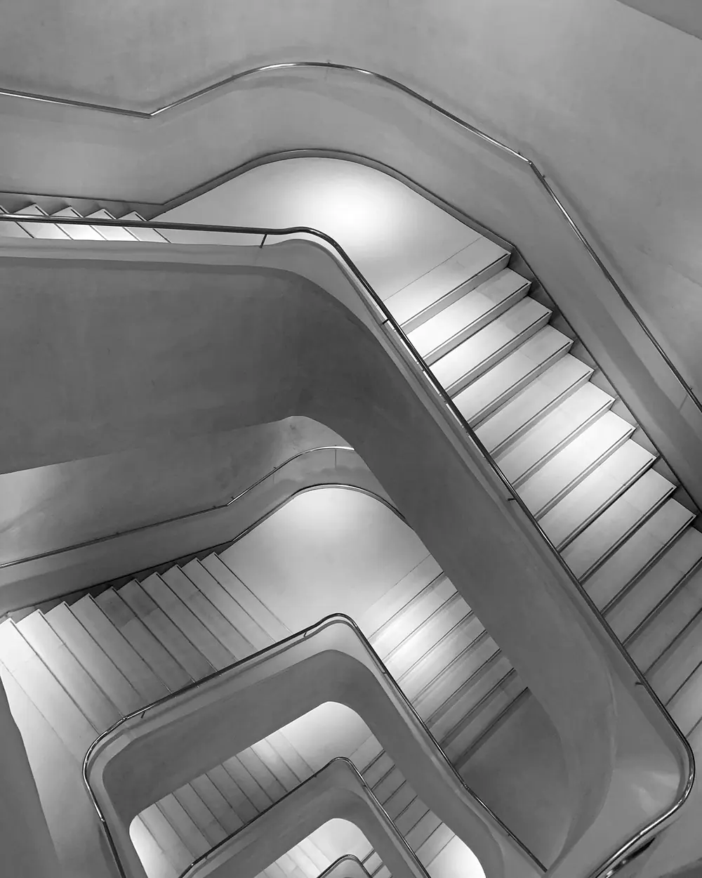 Un escalier de béton moderniste vu d’en haut, ses volées anguleuses se repliant les unes sur les autres.